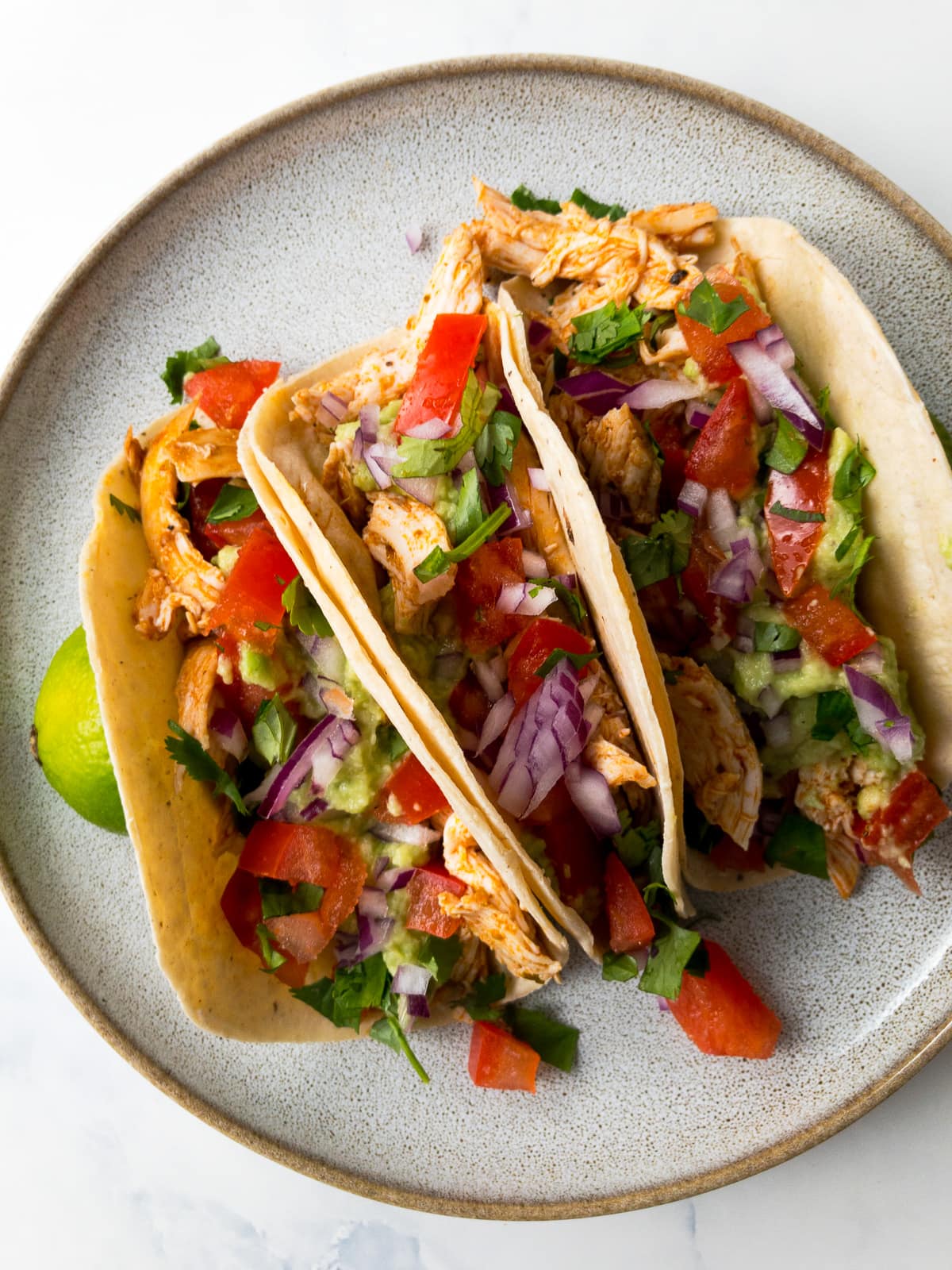 shredded chicken tacos topped with avocado, tomatoes, red onion, and cilantro on a plate with lime wedges