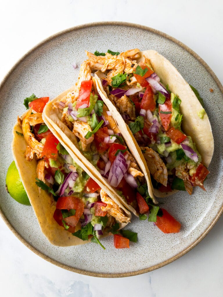 shredded chicken tacos topped with avocado, tomatoes, red onion, and cilantro on a plate with lime wedges