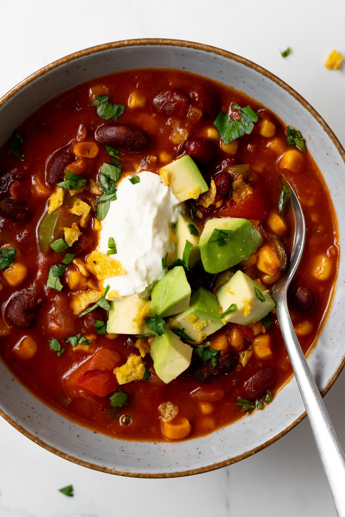 vegetarian chili garnished with avocado dices, sour cream, chopped cilantro, and tortilla chips in a bowl with a spoon