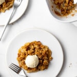 two plates of apple crisp with vanilla ice cream and two forks and a baking dish with apple crisp on the table