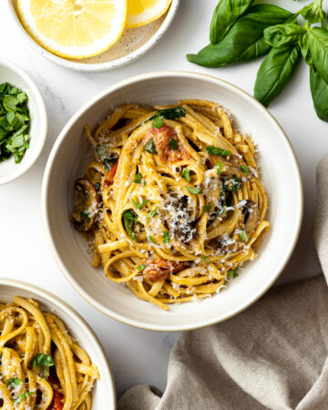 creamy pesto pasta in a bowl garnished with grated parmesan cheese and chopped fresh basil, another bowl of pasta, fresh basil leaves, and lemon slices on the same table