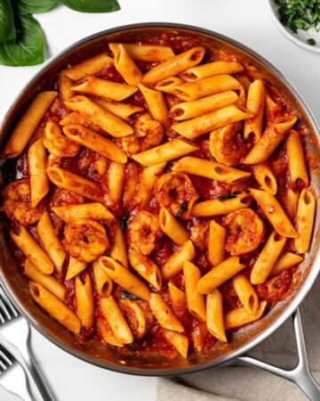 shrimp arrabbiata in a pan on a table and fresh basil, red pepper flakes, and two forks on the same table