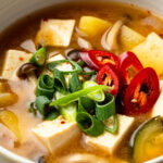 doenjang jjigae garnished with sliced green onion and sliced chili pepper in a bowl on a table