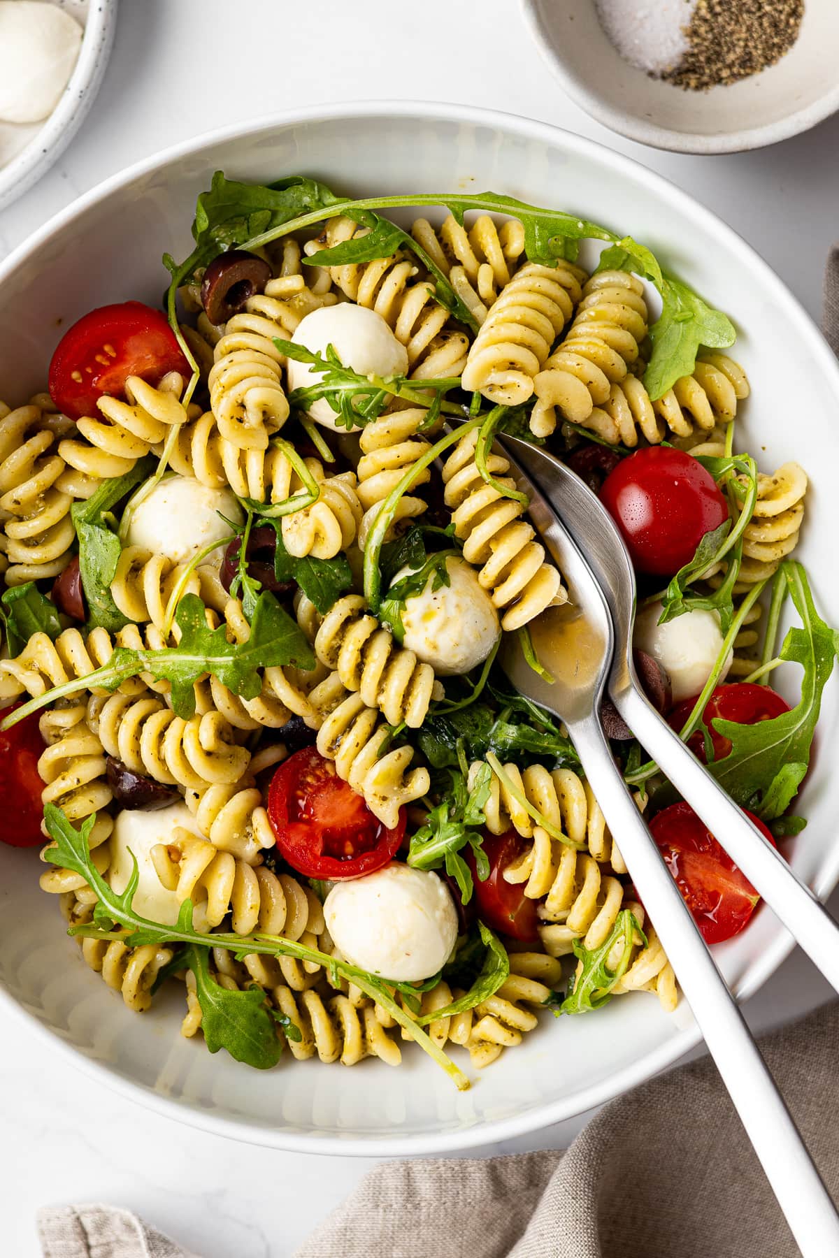 pesto caprese pasta salad in a bowl on a table with mozzarella pearls, arugula, and salt and pepper in separate bowl on the same table