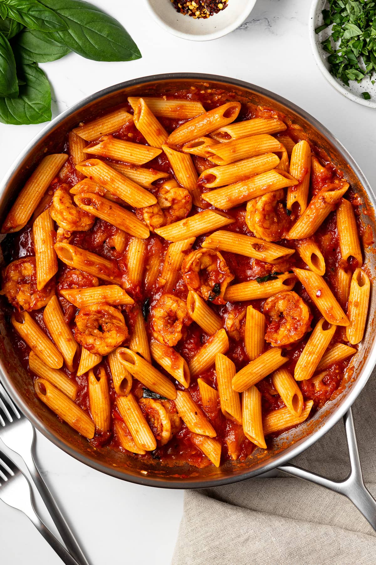 shrimp arrabbiata in a pan on a table and fresh basil, red pepper flakes, and two forks on the same table