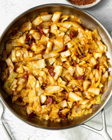 fried cabbage with bacon and onions in a pan on a table with a linen cloth, two spoons, and a bowl of Cajun seasoning on the same table