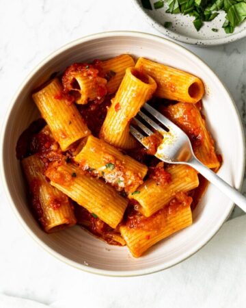rigatoni arrabbiata garnished with grated parmesan cheese and chopped basil leaves in a bowl with a fork