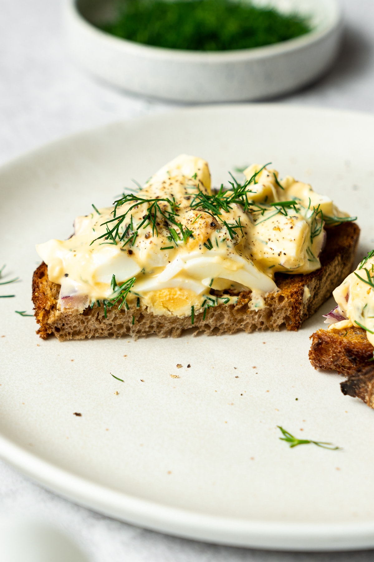 dill pickle egg salad garnished with chopped dill and black pepper on a slice of bread that is cut in half
