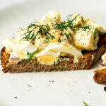 dill pickle egg salad garnished with chopped dill and black pepper on a slice of bread that is cut in half
