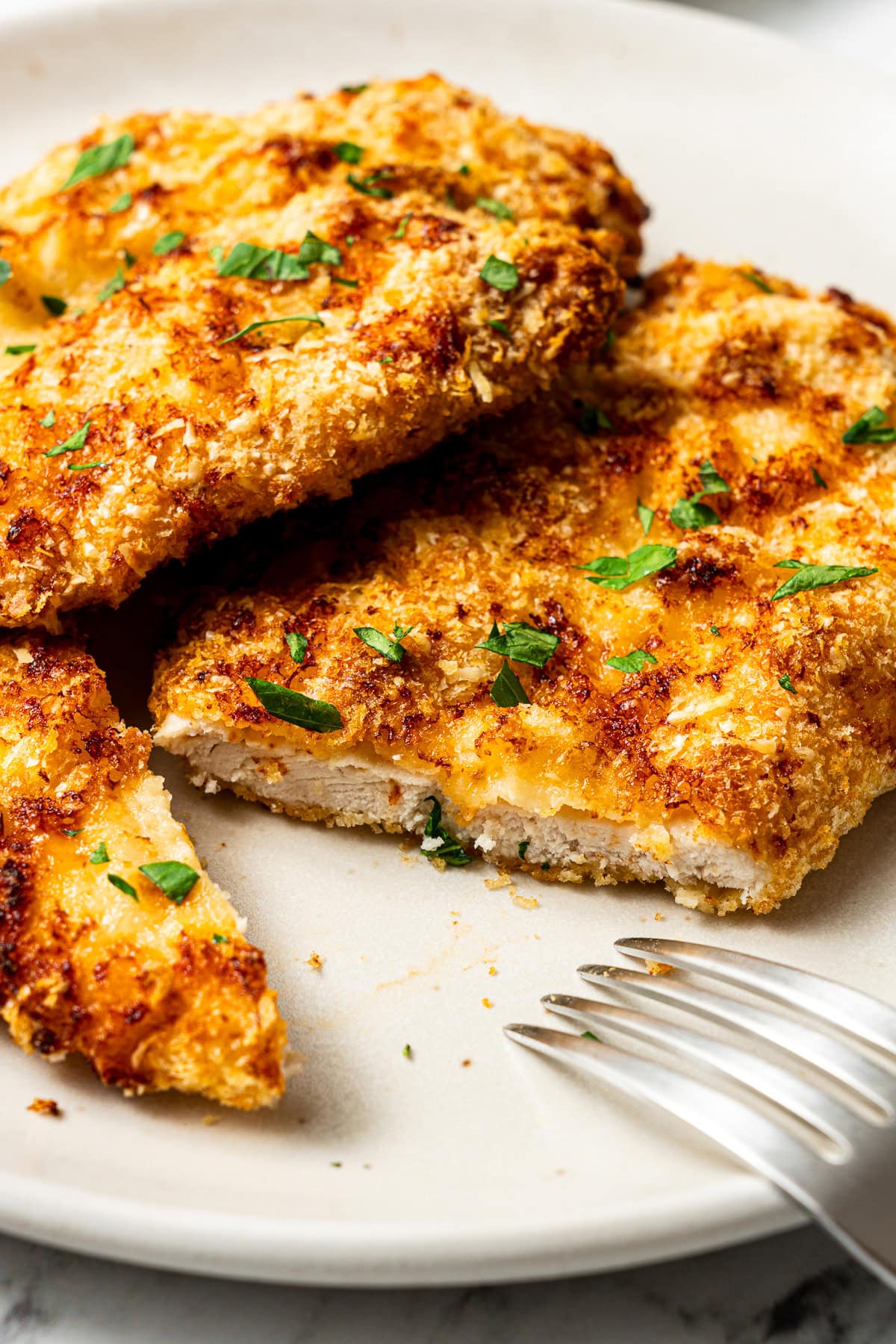 two air fryer parmesan crusted chicken cutlets on a plate garnished with chopped parsley; one cutlet is cut open