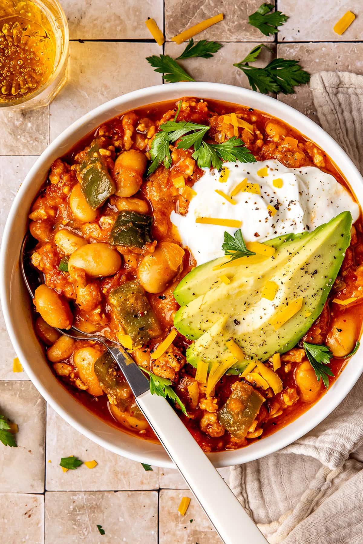 white bean pumpkin turkey chili in a bowl garnished with sour cream, cilantro, avocado, and shredded cheese with a spoon